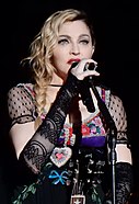 Madonna singing into a microphone over a black background, wearing a black lace dress with multi-coloured designs.