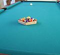 Racking up a game of seven-ball using a special hexagonal seven-ball rack, and incidentally also using a special 7 ball that borrows the black color and stripe, respectively, of the "money balls" in the games of eight-ball and nine-ball, to make it stand out more. The 1 ball is on the foot spot. (Expansive-view version.)