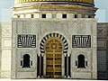 التركيز على الباب الرئيسي للضريح، أبواب خشب الساج تحمل النقوش التالية بالعربية "المقاتل الأسمى، باني تونس الحديثة، محرر المرأة" »
