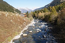 Il fiume scorre sulla largezza di 3 metri tumultuoso fra sterpaglie e vicino a una capanna di legno; sullo sfondo le montagne innevate delle Alpi.