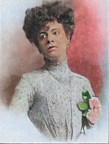 A colorized photo of Dina Alma De Paradeda; She's wearing a white dress, holding under her left arm, and staring into the distance.