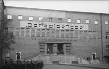 Grand bâtiment public avec inscriptions en allemand et une inscription plus grande qui imite les lettres de l'hébreu.