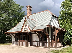 A white and brown wooden building with a central brick chimney, slate roof with copper trim and gutters, and tall, narrow windows with a brick surface in front and shrubbery behind it