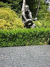 Horizontal gravestone saying "JAMES JOYCE", "NORA BARNACLE JOYCE", GEORGE JOYCE", and "…ASTA OSTERWALDER JO…", all with dates. Behind the stone is a green hedge and a seated statue of Joyce holding a book and pondering.