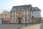 Photographie d'une place publique avec un bâtiment blanc de trois étages avec plusieurs fenêtres et un balcon central.