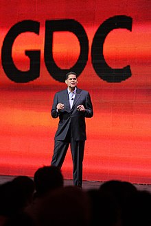 Homme se tenant debout sur une scène avec un fond rouge portant le sigle GDC.