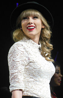 Taylor Swift wearing a brimmed hat and a laced white shirt smiling