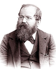 A grey scale image of a man aged between fifty and sixty-five looking to the left. He has a full length beard, oval glasses, his hair is ungroomed and he is going bald. He wears a three piece suit with the waistcoat buttoned up.