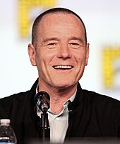 Мужчина средних лет с короткой стрижкой, в серой рубашке и черной куртке смеется в камеру с микрофоном, установленным перед ним.