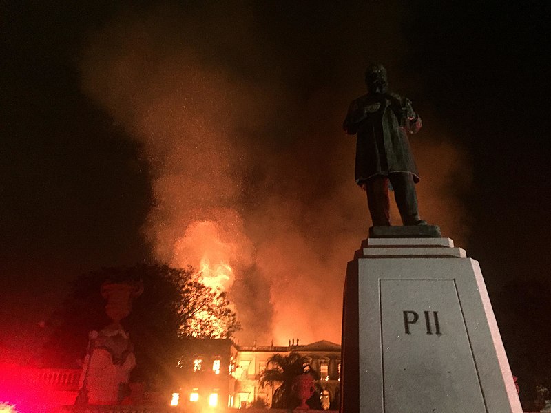 Photograph of a a large, tall building—the National Museum of Brazil—engulfed in flames; to the left of the image, the red glow of firetruck lights can be seen; and in the foreground, to the right, there is a statue of Emperor Pedro II, the name "Pedro II" engraved on its base