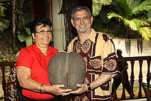 Larue schenkt Jorge Carlos Fonseca eine Nuss der Seychellenpalme, 2014. Eine Frau mit kurze Haaren und Roter Bluse überreicht die Melonengroße Nuss an einen Älteren Herrn mit einem Hemd mit traditionellen Mustern.