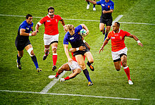 Farbige Obersicht auf ein Fußballfeld mit zwei blau gekleideten und drei rot gekleideten Spielern, die um einen Rugbyball kämpfen. Ein blau gekleideter Spieler klemmt sich den Ball unter seinen Arm und wird von einem rot gekleideten Spieler an den Hüften gepackt und versucht, ihn herunterzuziehen.