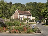 Au premier plan : vue d'un rond-point et de son terre-plein fleuri. Au second plan : vue d'un maison. À l'arrière-plan : les premiers arbres du bois de Sarron.