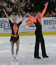 A figure skating couple wearing black and vivid orange clothing stands at the center of an indoor ice rink with their arms raised, thanking the audience; in the back, a portion of the crowd is visible.