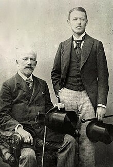 Two men in dark jackets, waistcoats and light slacks and carrying top hats and canes, a man in his 20s with dark hair standing and the second in late middle age with balding grey hair and a beard