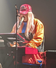 Tones and I is standing behind a keyboard while also singing into a microphone. She wears red and white cap with the lettering "The Kids Are Coming". She also wears a multi-coloured jacket and is looking down at a sheet on a stand.