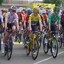 Cyclists in polka-dot, yellow and green jerseys ride side-by-side, leading the peloton