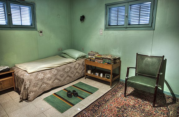 Photograph of Ben-Gurion's hut, depicting a modest abode with a thin bed in the corner, a low bookcase to the right of it and a low cabinet to its bottom, a single chair further right atop a rug