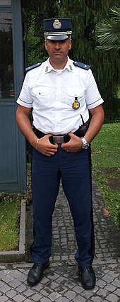 Frontale Farbfotografie von einem Mann in Gendarmenuniform. Er trägt eine dunkelblaue Mütze mit Emblem, ein weißes kurzärmeliges Hemd mit zwei Knöpfen auf den Schulterklappen, eine dunkelblaue Hose und schwarze Schuhe. An seiner linken Brust ist ein Ansteckschild.