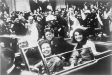 Photo noir et blanc de quatre personnes souriantes à bord d'une voiture décapotée, traversant une foule enthousiaste.