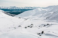 "A top-down view of the site, showing approximately 10 wooden buildings in the snow. Groomed cross-country ski trails weave through and around the buildings."