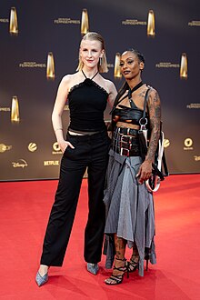 Zwei Frauen, links mit heller und rechts mit dunkler Hautfarbe in Abendgarderobe vor Sponsorenwand