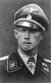 A black-and-white photograph of a man wearing a military uniform, peaked cap and a neck order in shape of an Iron Cross. His cap has an emblem in shape of a human skull and crossed bones. A large scar on his chin is visible.