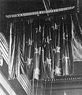 Черно-белое фото потрепанного в боях флага, подвешенного к потолку музея.