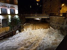 Photo de nuit montrant la Durolle en crue avec un très gros débit d'eau brunâtre au niveau d'une petite cascade en pleine ville, avec une usine sur la rive, à gauche du cliché