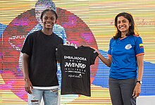 Linda Caicedo holding the left sleeve of a black t-shirt that reads "Embassador of the Resilient Youth." Anu Rajaraman is to her right, holding the right sleeve of the t-shirt.