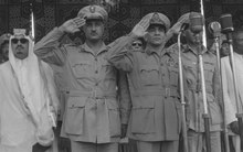 Five men in military uniforms standing in a row, with the three in the middle saluting