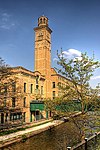 A three-story pale-red brick building along the left side of a small river. The building has a bell tower rising out of the middle of it.