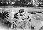 Black and white photo of two women sitting in a hammock in turn of the 20th century dresses; one reclines and the other sits on her lap and wraps her arm around the other, both staring at each other.