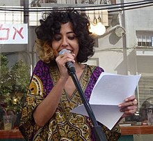 Keissar on stage, speaking into a microphone. The image is of the upper half of her body. She is wearing a purple and gold traditional-Yemeni style top. She has dark curly hair, and is holding several sheets of paper in her left hand.