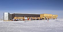 A long, large building consisting of several sections stands behind a line of flags flying on poles. The ground surface is ice-covered; in the middle foreground is a short striped pole which indicates the position of the South Pole