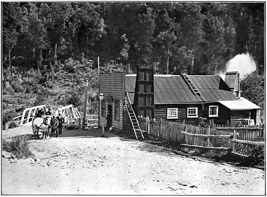 House with dirt road in front and a picket fence to the right, a horse and dray in the front with men around it, a bridge behind them, with trees on the slopping hill in the background