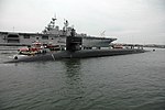 Submarine in port with an aircraft carrier in the background.