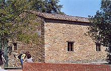 Photo of a building of rough stone with small windows, surrounded by olive trees.