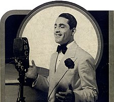 A vintage photo of Al Bowlly wearing a tuxedo and singing into a NBC microphone.