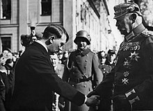 Photographie en noir et blanc prise à Potsdam le 21 mars 1933. À gauche de l'image, Adolf Hitler, vêtu d'une redingote, serre la main du président von Hindenburg, en s'inclinant vers lui. À droite, Hindenburg domine Hitler d'une bonne tête et se tient droit. Ganté, il porte le grand uniforme de maréchal prussien, arborant de nombreuses décorations dont la croix de fer, et un casque à pointe. Au centre et à l'arrière plan, entre les deux hommes, un militaire avec le lourd casque de combat de l'armée allemande