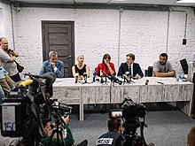 Cinq personnes installées à une longue table blanche font face à des journalistes.