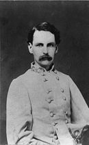 Black and white photo shows a mustachioed man with dark hair. He wears a gray military uniform with the left sleeve empty.