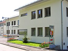 Gebäude mit heller Fassade, davor auf einer Glasstele das Wappen der Gemeinde, darüber die Aufschrift "Gemeindeamt St. Gotthard", darunter "Hort" (für die Kinderbetreuungsstätte im Untergeschoß des Gebäudes)