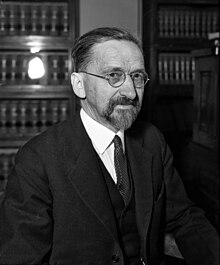 Photograph of a siting Caucasian male from the waist up wearing glasses and a three-piece suit. He has a mustache and short beard.