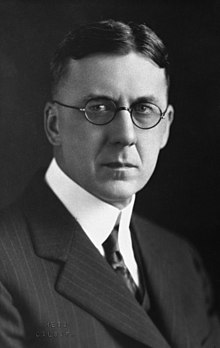 A severe-looking man wearing round-rimmed glasses