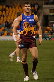 A man in a maroon, blue and gold Australian rules football jersey walks towards the camera on grass