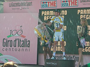 A cyclist popping a champagne bottle on a podium, flanked by two women holding flowers and wine.