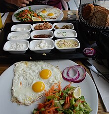 Plates with fried eggs, green salad, and onions; basket of bread; smaller dishes filled with variety of sides.