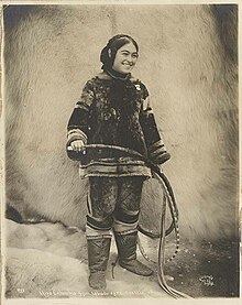 A smiling young Inuit woman, wearing traditional clothing, holding a whip