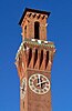 A narrow square brick tower. At its bottom is a clock with white Roman numerals. Above it is a balcony with narrowly spaced brick supports; on top is a narrower section with a narrow round-topped opening and an overhanging roof.